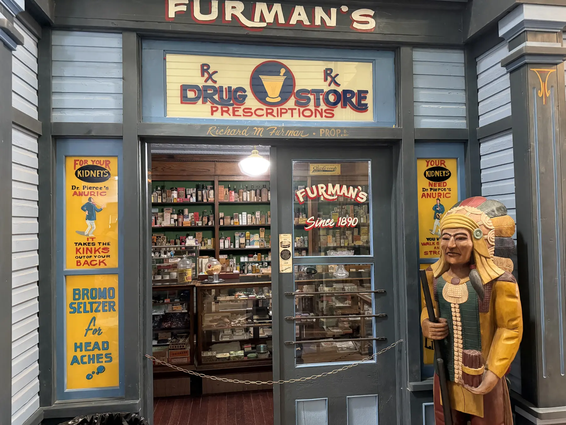 A reconstructed drugstore storefront with painted signs, shelves, and a carved wooden figure