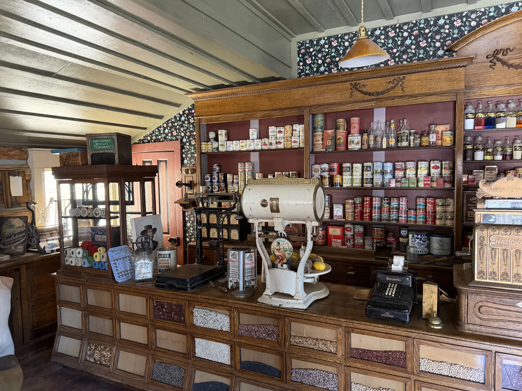 Shelves of period groceries, a scale, coffee grinder, and cash register inside the general store