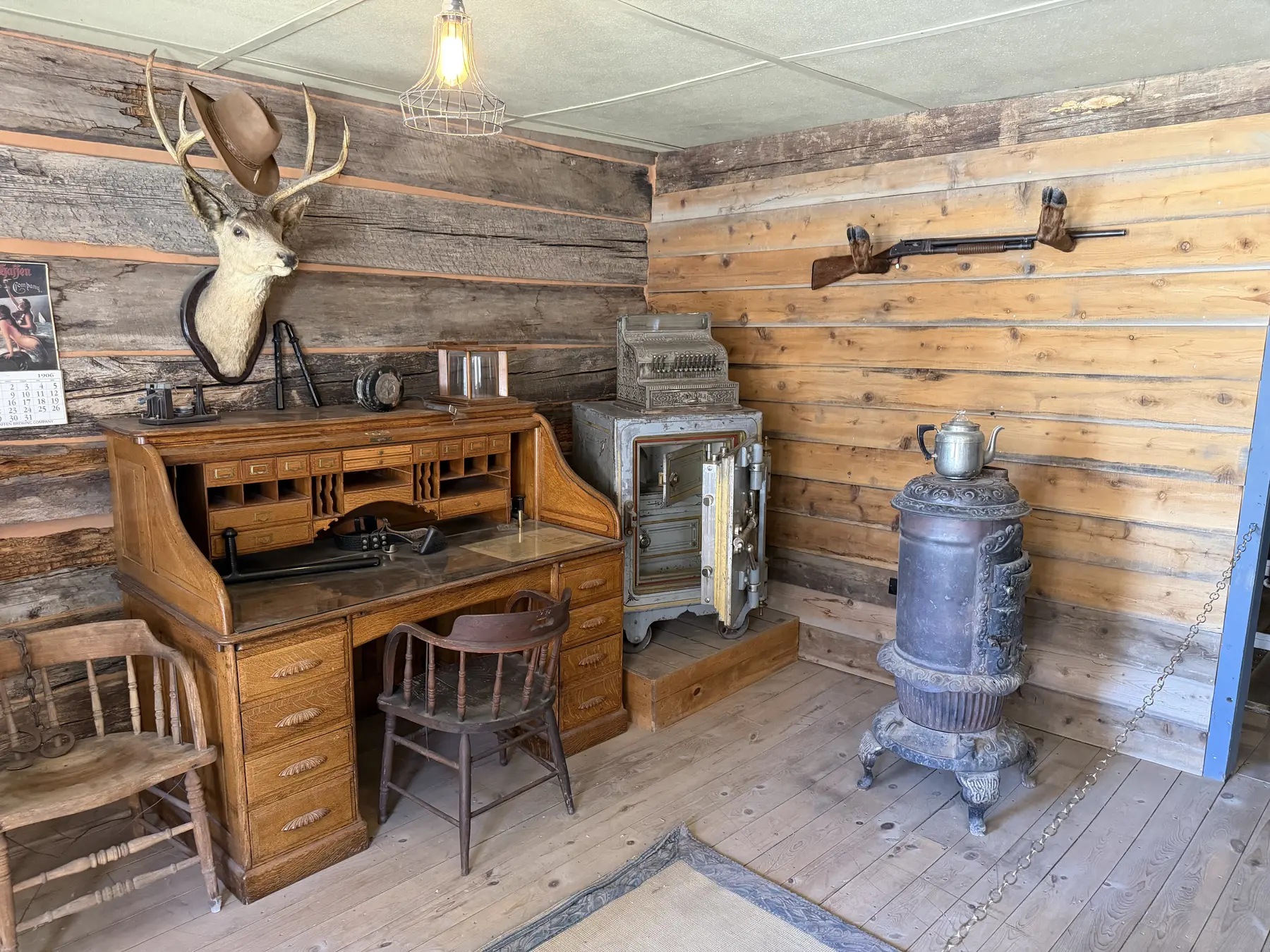 A furnished log-cabin room with a wooden desk, chair, stove, and mounted deer