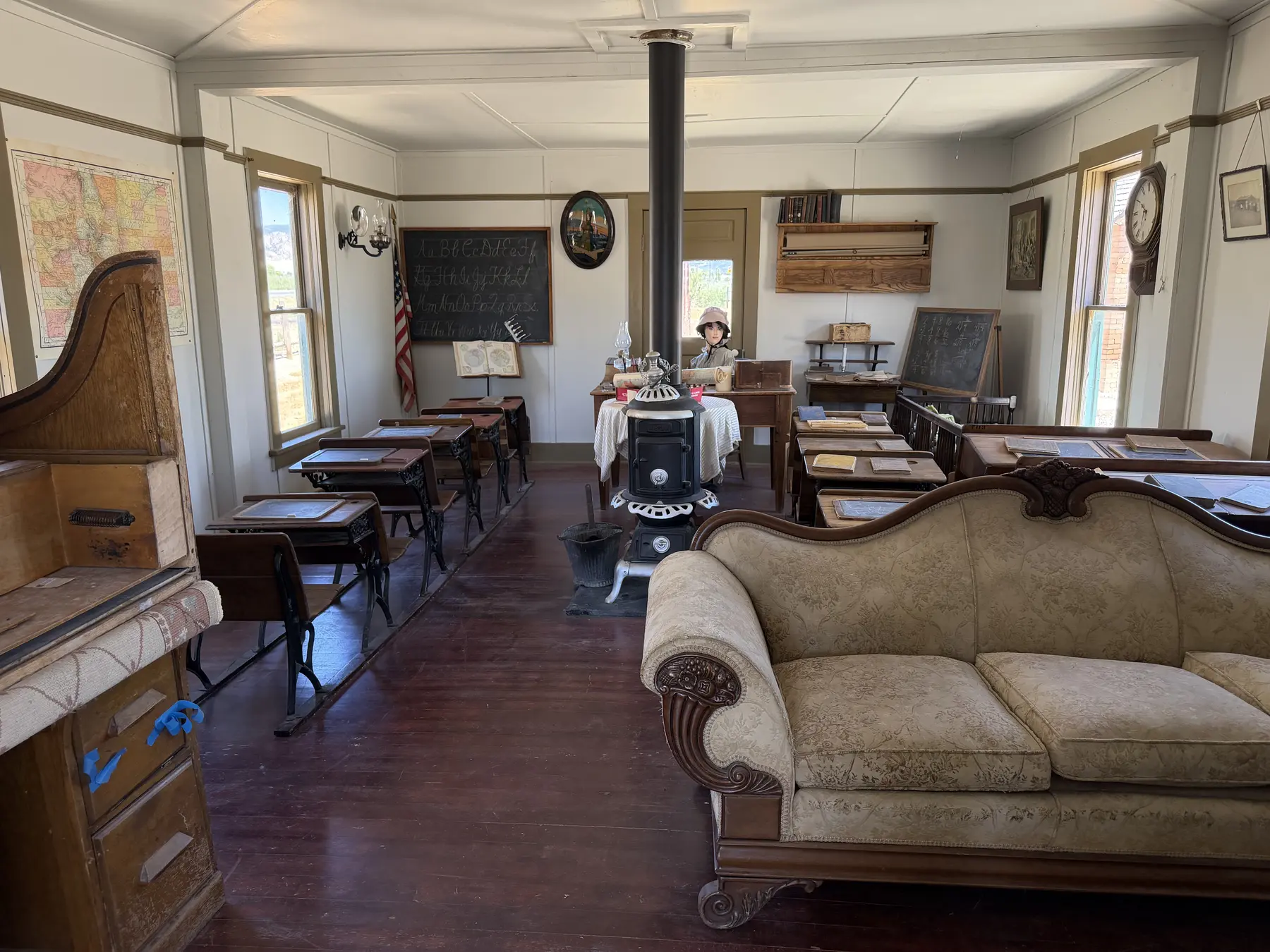 A historic schoolroom with desks, maps, a stove, piano, and teacher's table